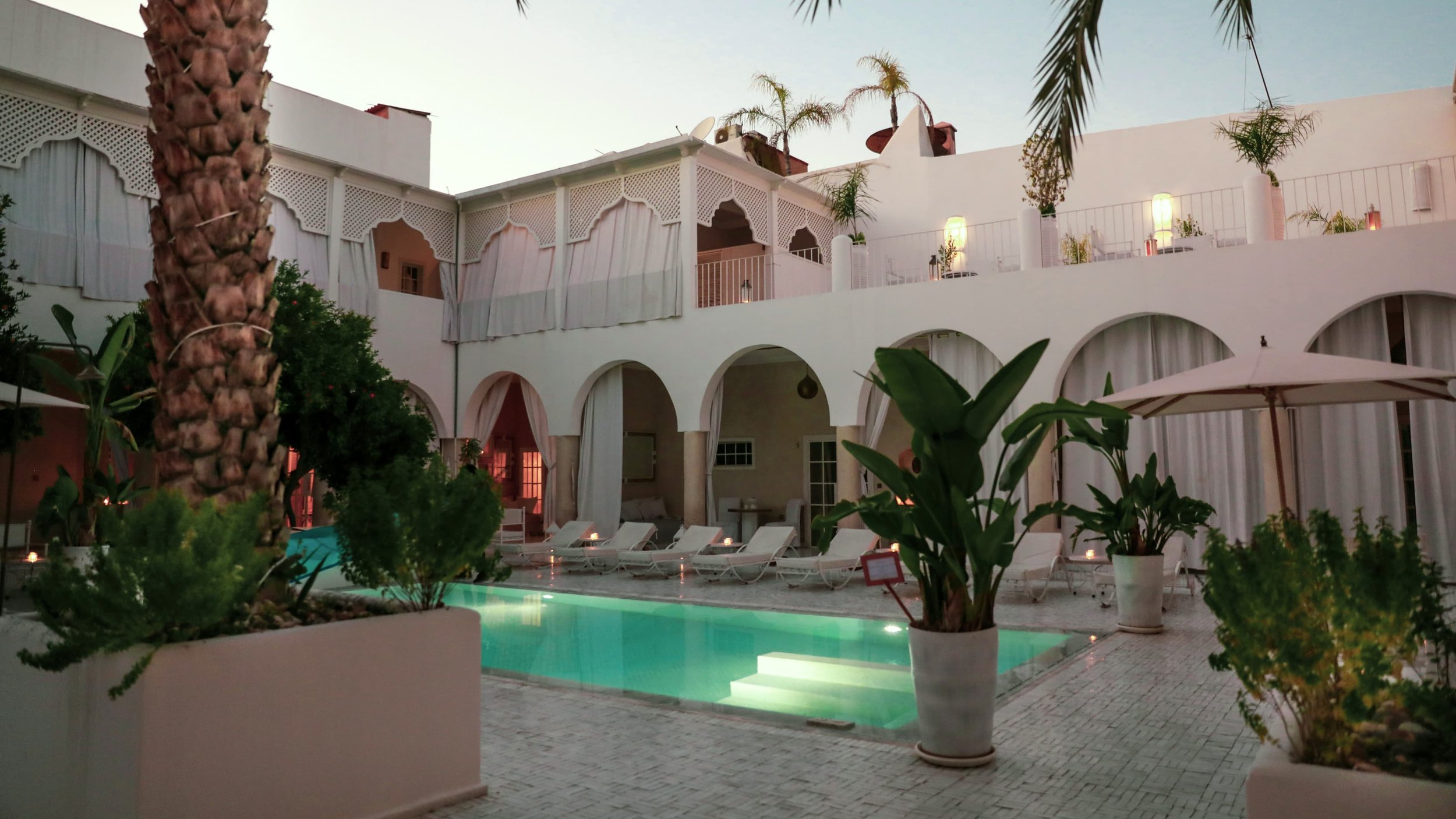 Cour de riad au crépuscule : bassin éclairé, arcades blanches et palmiers