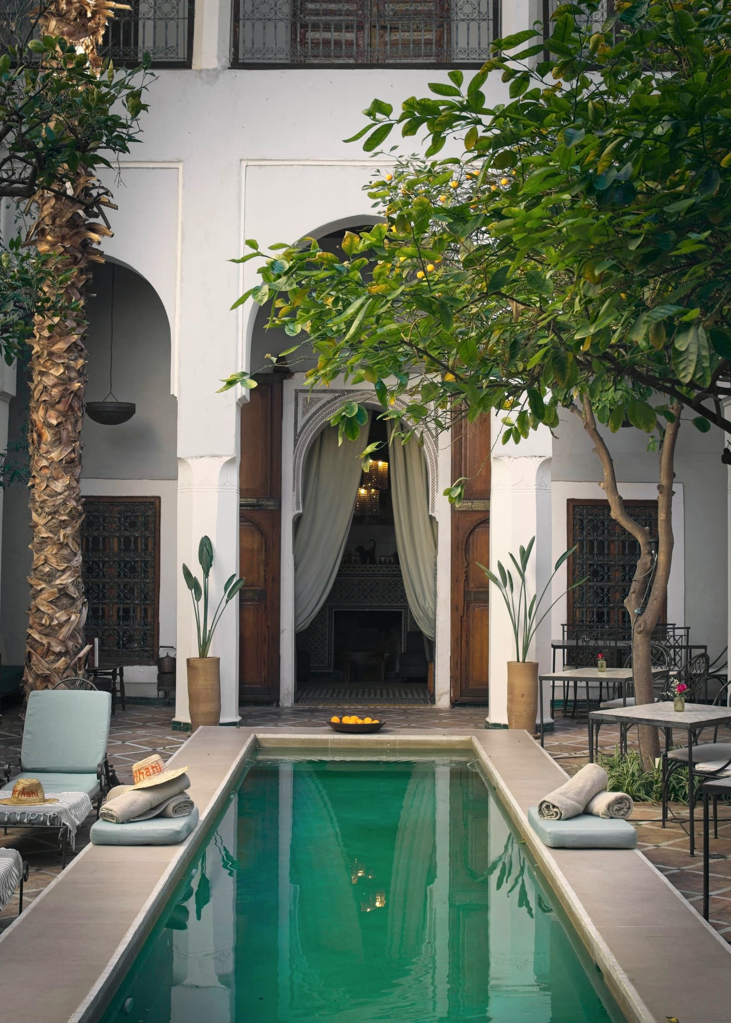 Patio de riad avec long bassin, arcades, palmier et rideaux de lin
