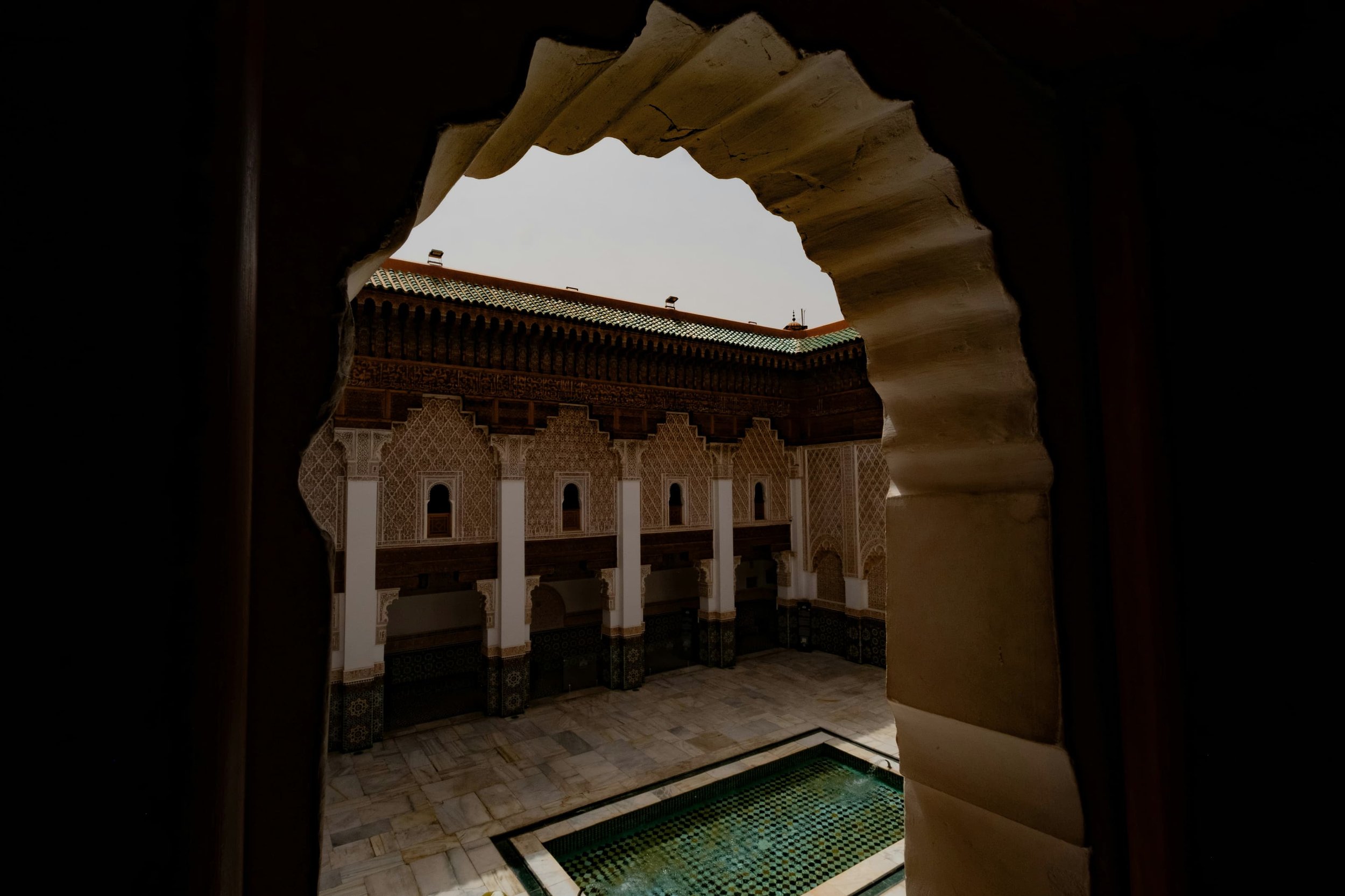 Courtyard with carved arcades and a zellige pool, seen through a scalloped arch